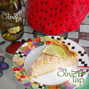 A slice of Persian lime custard tart topped with toasted coconut and a lime wedge, served on a colorful dessert plate with a bottle of olive oil and a decorative strawberry in the background.