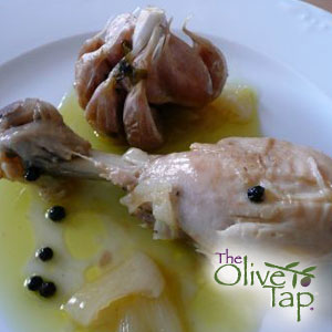 A chicken drumstick next to a roasted bulb of garlic with a yellow sauce and capers on a white dish.