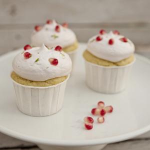 Three small cakes with white wrappers topped with whipped cream and pomegranate seeds, served on a white cake stand.