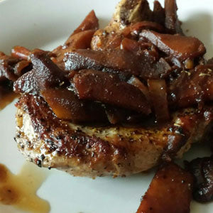 A seared porkchop topped with sauteed apples and onions, served with pan sauce on a white plate.