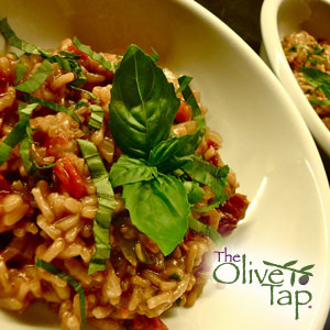Risotto with sundried tomatoes garnished with chopped basil and a fresh basil leaf served in a white bowl with The Olive Tap logo in the bottom righthand corner.