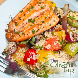 A roasted trout fillet served over couscous with roasted vegetables, including peppers, onions, and zucchini, garnished with chopped parsley on a white plate.