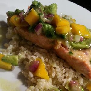 Riced cauliflower topped with baked salmon and an avocado, red onion, and mango salsa, served on a white plate.