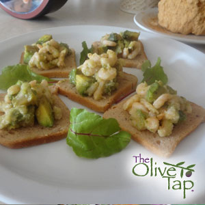 Toasted appetizer bread topped with avocado and shrimp salad, served on a white plate with leafy greens.