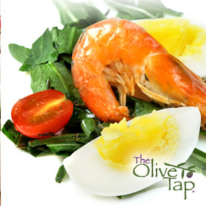 A shrimp prawn on a bed of spring mix salad with quartered hard-boiled eggs and a half of a cherry tomato on a white surface.