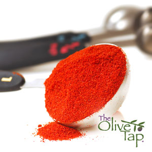 A close-up of a measuring spoon with smoked paprika on a white surface with measuring spoons in the background with The Olive Tap logo in the bottom righthand corner.