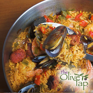 A paella with sausage, mussels, shrimp, and roasted red peppers in a metal bowl on a wooden background.