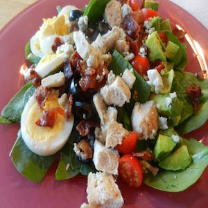 Spinach cobb salad topped with cherry tomatoes, hardboiled eggs, avocado chunks, bacon crumbles, and croutons, served on a pink plate.