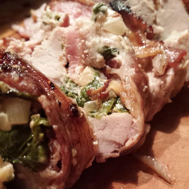 Sliced pork tenderloin stuffed with spinach, goat cheese, and bacon, served on a wooden cutting board.