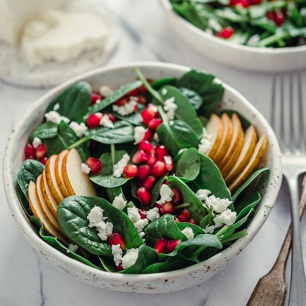 Spinach salad with sliced pears, pomegranate seeds, and crumbled feta cheese served in a white bowl.