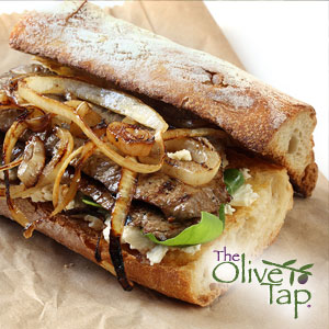 Steak on a toasted bread loaf with grilled onions, arugula, and gorgonzola cheese crumbles on a brown parchment paper with a white background.