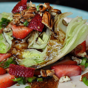 A crisp wedge of iceberg lettuce topped with sliced strawberries, walnuts, green onions, and feta cheese crumbles, drizzled with balsamic vinegar.