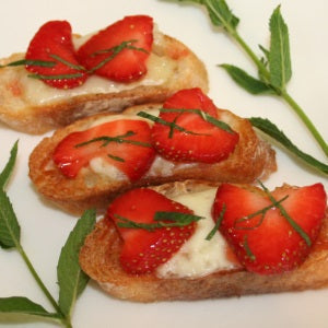 Three toasted crostini topped with brie cheese, fresh sliced strawberries, and finely chopped mint, arranged on a white plate with mint sprigs.
