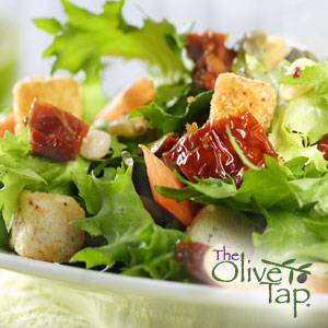 A close-up of lettuce with croutons, sundried tomatoes, feta cheese, and pine nuts with The Olive Tap in the bottom righthand corner.