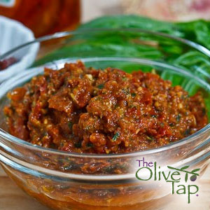 A red sundried tomato pesto in a glass bowl on a wooden surface with The Olive Tap logo in the bottom righthand corner.