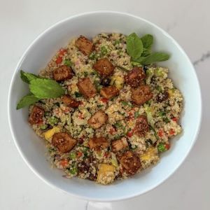 A bowl of quinoa salad with teriyaki tofu cubes, mango, and garnished with fresh mint leaves in a white bowl.
