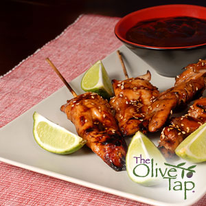 Grilled, glazed chicken skewers with a side of dipping sauce, served on a white plate with lime wedges.