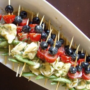 Tortellini skewers with black olives, grape tomatoes, and snap peas drizzled with a dill vinaigrette and arranged on a serving platter.