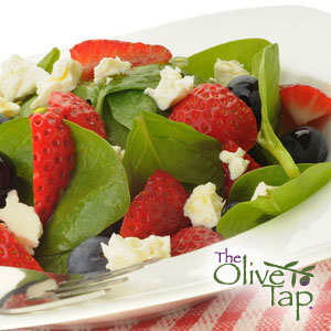 Spinach, cut strawberries, blueberries, and feta cheese crumbles in a white bowl on a picnic tablecloth.