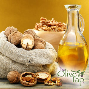 A glass pitcher halfway full of walnut vinaigrette next to a bag of whole walnuts, a few cracked open, with a bowl of shelled walnuts in the background, all against a honey-colored background and on a gray surface.
