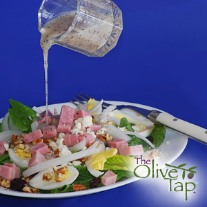 A white plate with a leafy green mixture with romaine topped with cubed ham and hard-boiled egg slices, being drizzled with a honey mustard vinaigrette from a glass cup, a white fork sitting on the plate, all against a dark blue background.