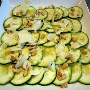 Slices of zucchini topped with pine nut0s and shaved parmesan cheese on a white plate.