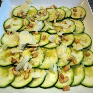 Slices of zucchini topped with pine nut0s and shaved parmesan cheese on a white plate.