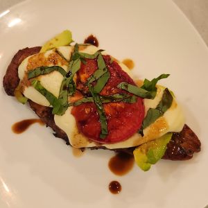 Chicken breast topped with avocado, mozzarella cheese, tomato slice, and chopped fresh basil drizzled with balsamic vinegar on a white plate.