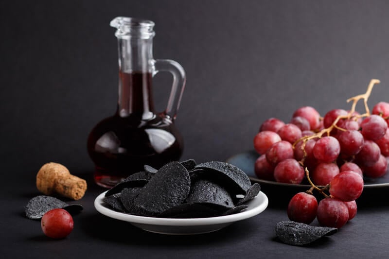 A glass cruet filled with dark balsamic vinegar, displayed beside a plate of red grapes and black potato chips on a dark background.