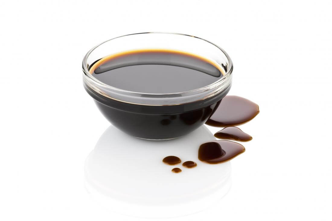 A small glass bowl of balsamic vinegar with a few drops on the side on a white background.