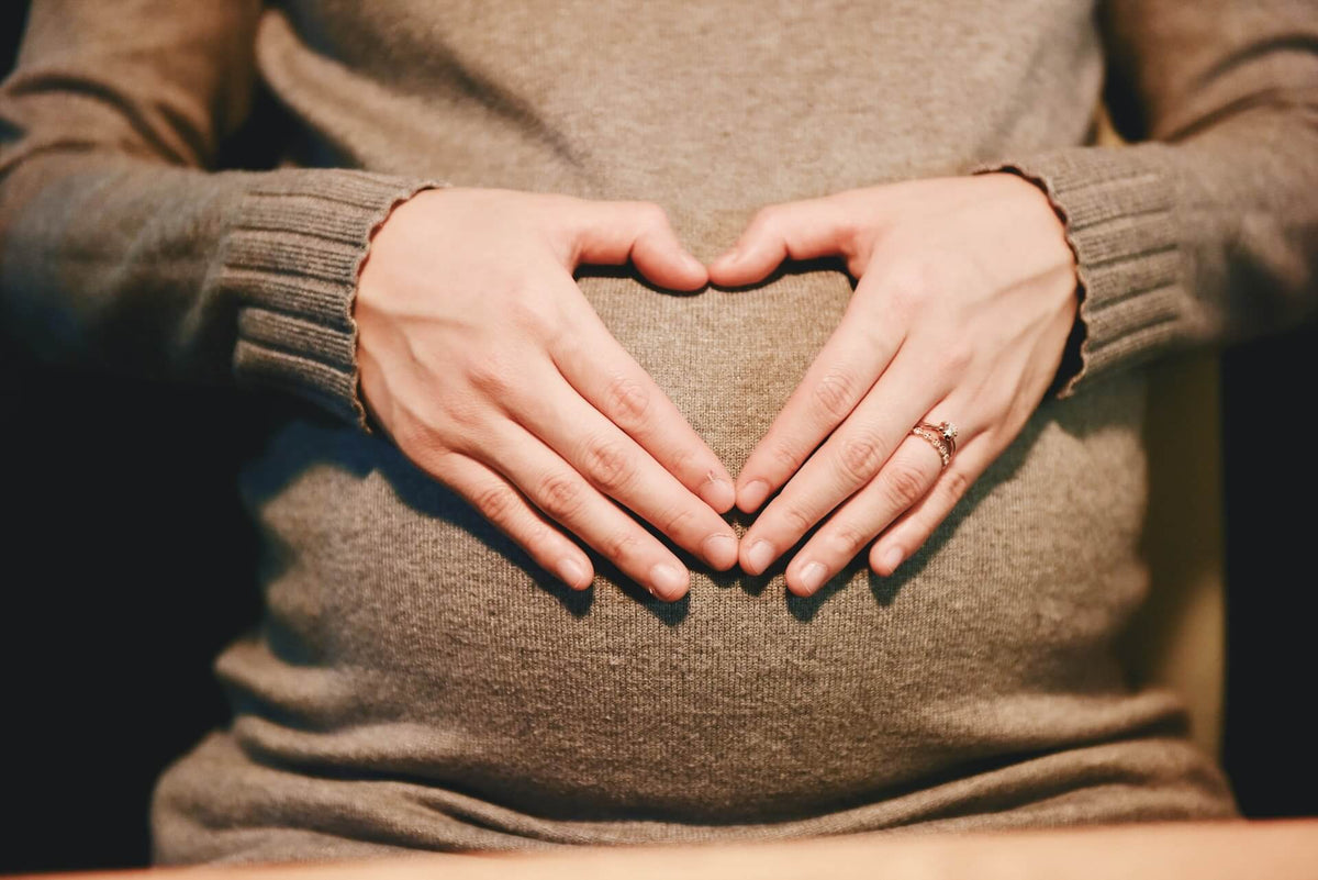 A person wearing a knit sweater with their hands forming a heart shape over a pregnant belly.