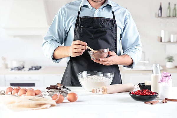 A person in an apron preparing ingredients at a kitchen counter, with a mixing bowl, eggs, flour, milk, and baking supplies nearby.