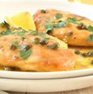 Two breaded chicken breasts topped with capers and herbs, served in a lemon caper sauce with lemon wedges on a white plate.