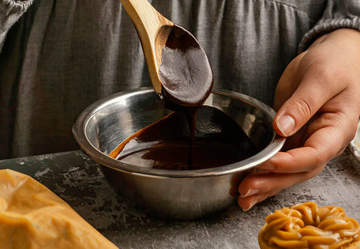A wooden spoon drizzling chocolate balsamic glaze into a metal mixing bowl, held by a person preparing a sauce.