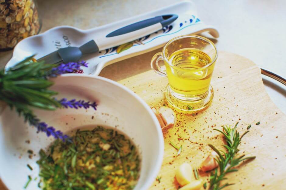 A small glass of olive oil on a wooden cutting board next to fresh rosemary, garlic, and a bowl of olive oil mixed with fresh herbs with fresh lavender on the bowl.