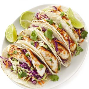 Fish tacos filled with grilled white fish, shredded red and green cabbage, carrots, and fresh cilantro, served on soft tortillas with lime wedges on a white plate.