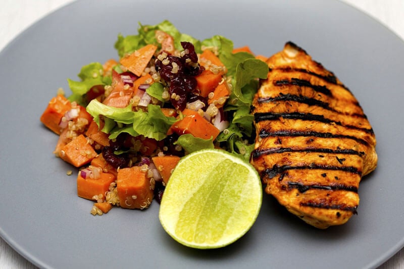 Grilled chicken breast with quinoa, sweet potato, and cranberry salad served with fresh lime on a gray plate.