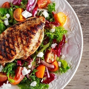 A grilled chicken breast on mixed greens with feta cheese crumbles, tomatoes, and red onion on a white plate on a wooden surface.