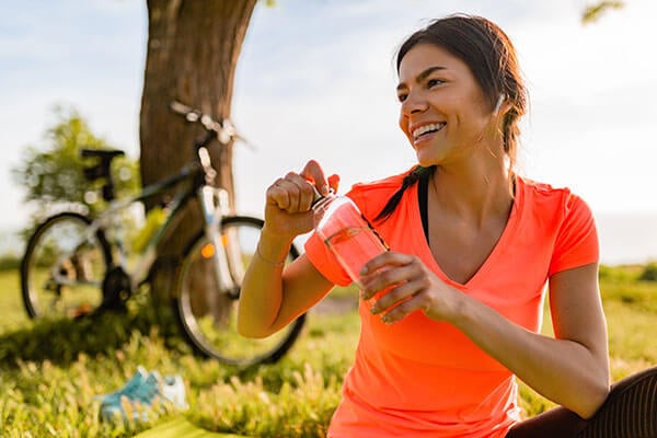 A woman holding a water bottle while sitting outdoors, with a bicycle in the background leaning against a tree.