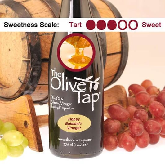 The Olive Tap's Honey Balsamic Vinegar with a picture of fresh honey, surrounded by grapes in front of a wooden barrel, and a sweetness scale indicating a three on a light background.