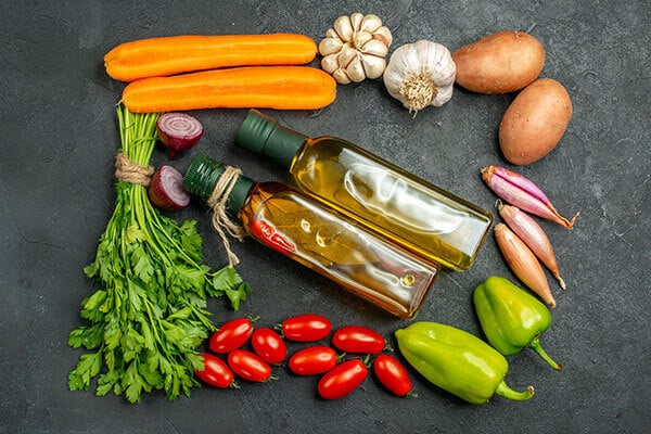 Two bottles of olive oil surrounded by parsley, cherry tomatoes, peppers, shallots, sweet potatoes, garlic, a carrot, and red onion all making a rectangular shape around the bottles on a black surface.