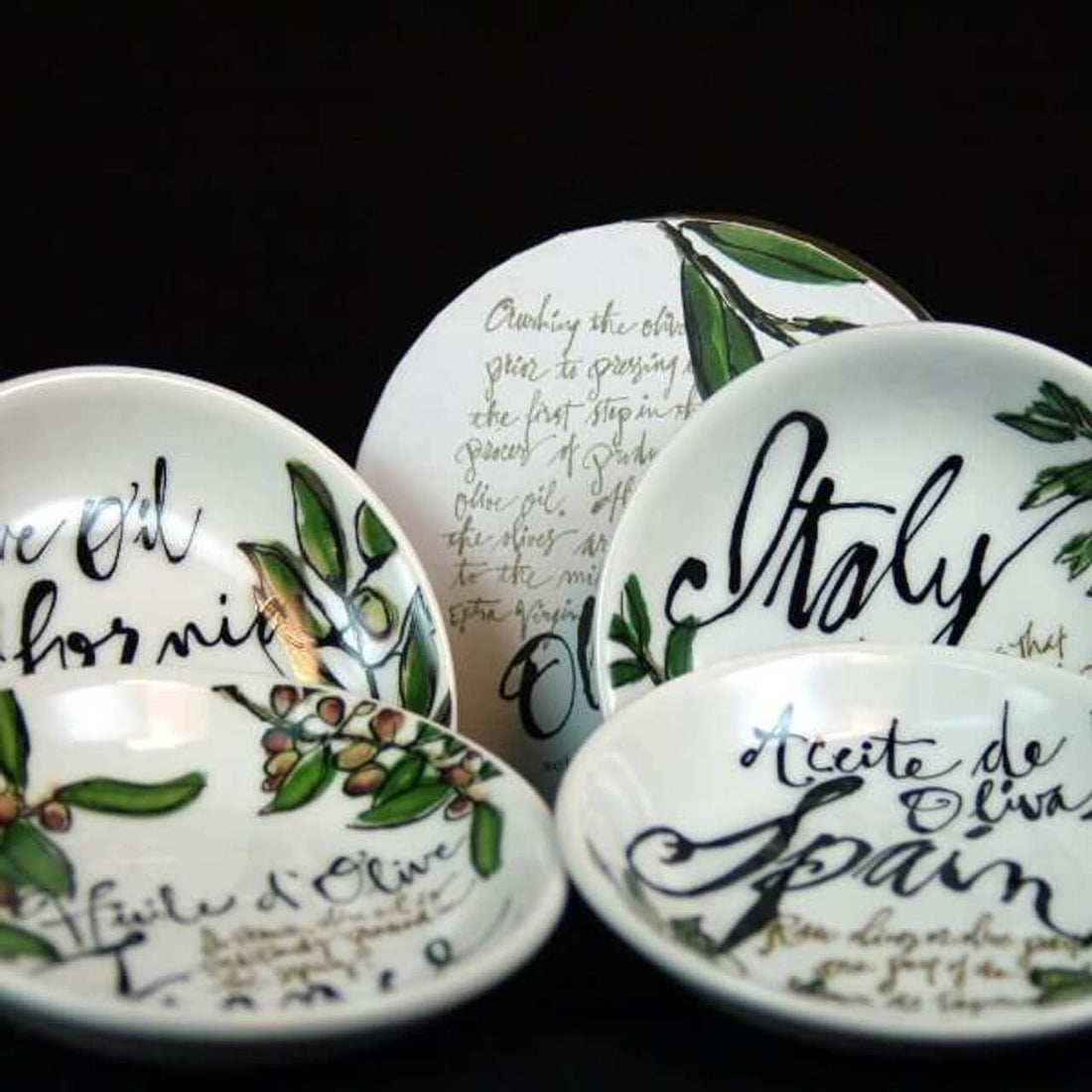A collection of ceramic dishes decorated with olive branches and olive oil-themed lettering, displayed against a black background.