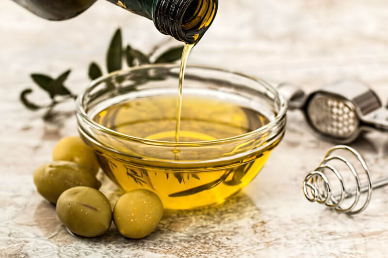 Olive oil being poured into a small glass bowl, surrounded by olives, a garlic press, and whisk on a light-colored surface.