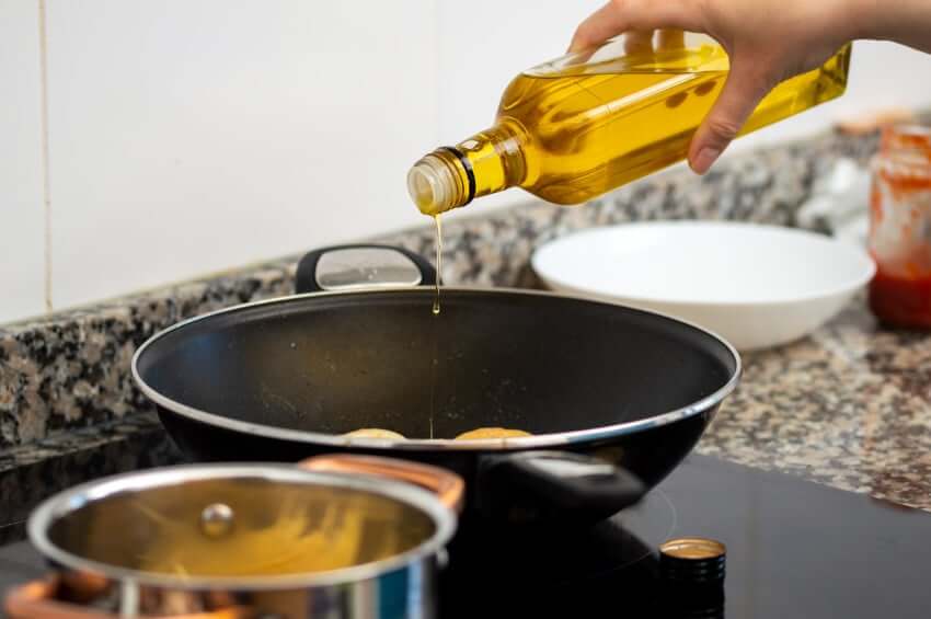 A hand pouring olive oil from a glass bottle into a frying pan on a stovetop, with cookware and kitchen items nearby.