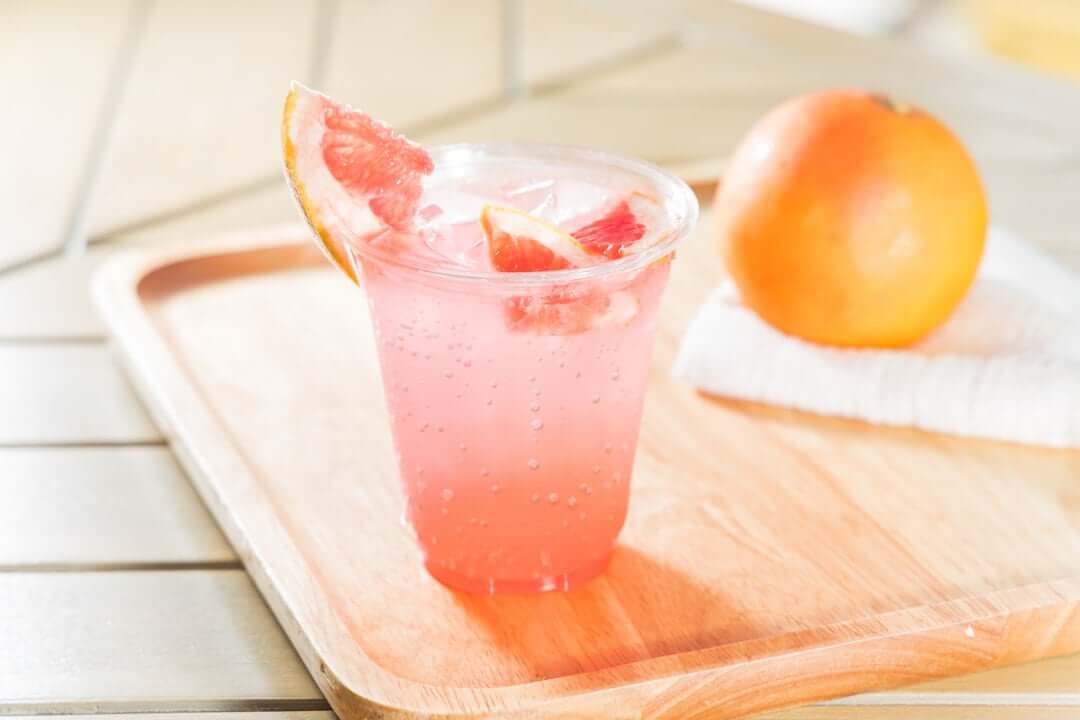 A plastic cup with pink liquid garnished with a slice of grapefruit on a wooden cutting board with an orange in the background on a towel.