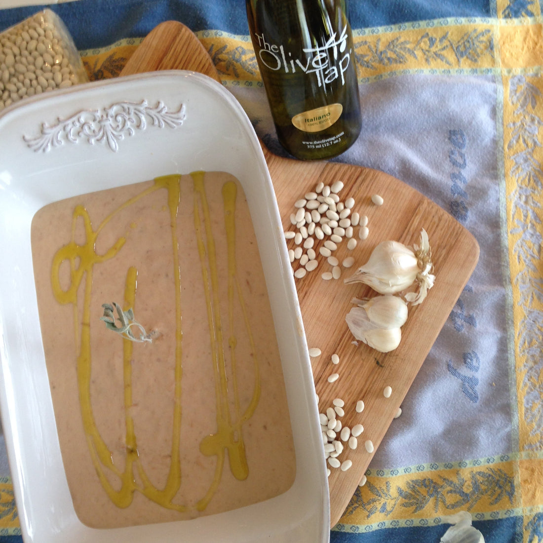 Creamy white bean puree drizzled with olive oil in a baking dish, served alongside garlic gloves and white beans on a wooden board.
