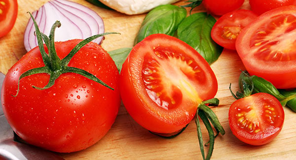 Fresh whole and sliced tomatoes with basil leaves, red onion, and mozzarella cheese arranged on a wooden surface.