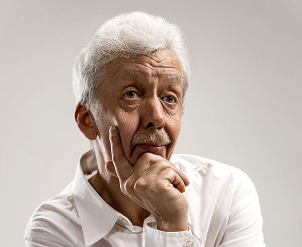 An older man with white hair wearing a white shirt, resting his chin on his hand with a thoughtful expression.