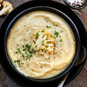 White creamy cauliflower soup garnished with a piece of cauliflower and chopped parsley in a black bowl.