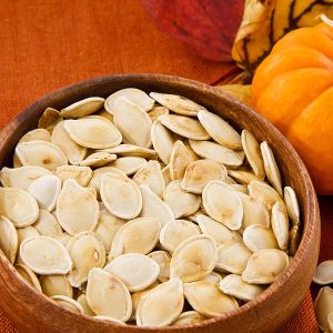 Roasted pumpkin seeds in a wooden bowl next to a pumpkin and a fall background.